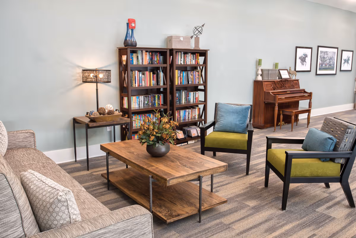A cozy living room area with a gray sofa, two green cushioned chairs with blue pillows, a wooden coffee table with a flower arrangement, a side table with decorative items and a lamp, two tall bookshelves filled with books, and a wooden piano against a light blue wall adorned with framed pictures.