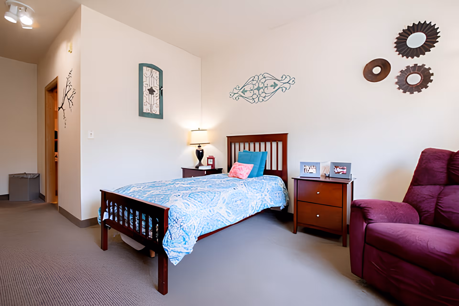 Simple bedroom with a single wooden bed, nightstand and lamp, wall decorations and a purple recliner.