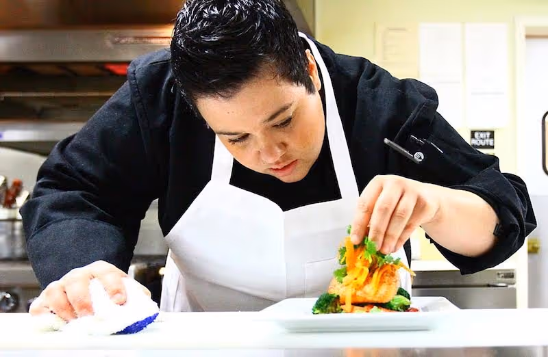 Chef in a white apron carefully plates a dish in a commercial kitchen.