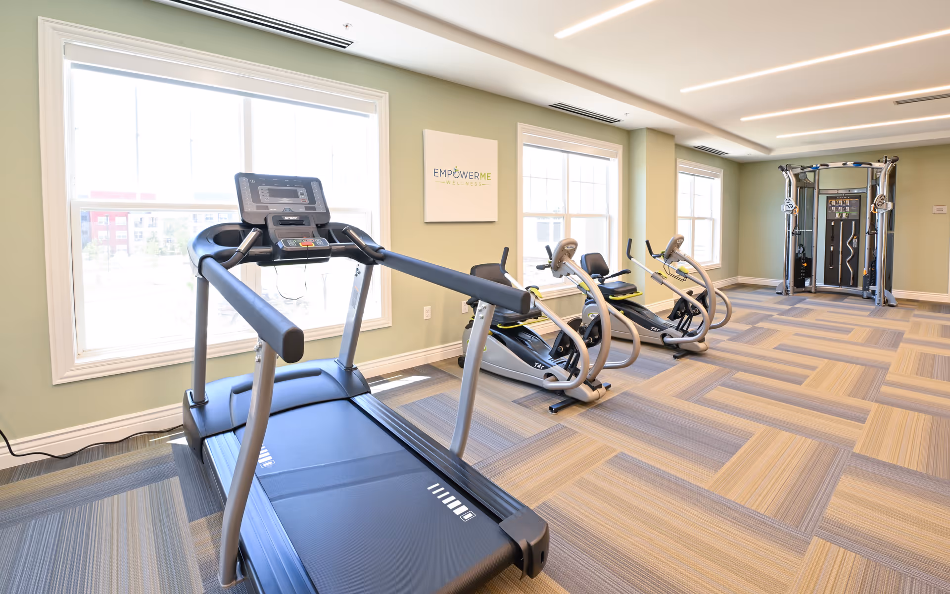 A bright fitness room with large windows letting in natural light. The room contains a treadmill, two recumbent exercise bikes, and a multi-functional weight machine. The walls are painted light green and there is a sign on the wall that reads 'EMPOWER ME WELLNESS'. The floor is covered with patterned carpet tiles in shades of beige and gray.