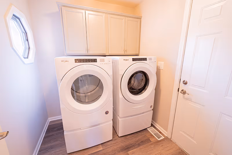 A laundry room with a white front-loading washer and dryer set side by side. Above the appliances are beige cabinets for storage. The room has light-colored walls, a small octagonal window on the left wall, and a white door on the right side. The floor is covered with wood-style vinyl or laminate.