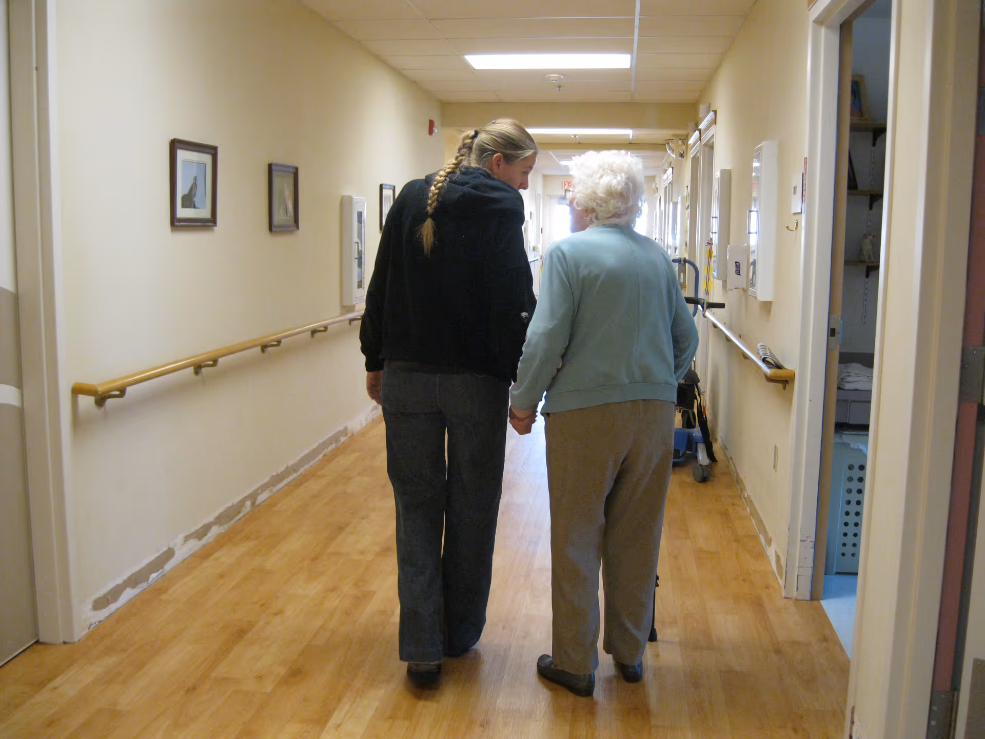 A younger woman with a braid is walking down a hallway holding hands with an elderly woman using a walker. The hallway has wooden floors, handrails on both sides, and framed pictures on the walls.