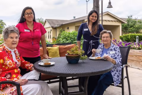 Two elderly women sitting at a round outdoor table with coffee cups and snacks, accompanied by two staff members standing behind them. The setting is a patio area with greenery and a building in the background under a cloudy sky.