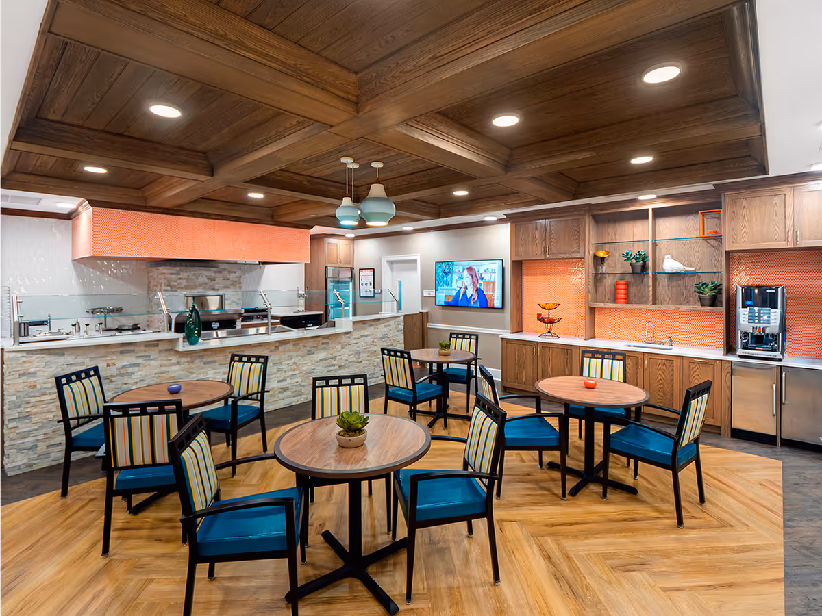 A bright and modern dining area with round wooden tables and chairs featuring blue cushions and striped backs. The room has a wooden coffered ceiling with recessed lighting. There is a serving counter with a stone facade and a kitchen area behind it, including cabinets, a coffee machine, and decorative shelves with plants and ornaments. A flat-screen TV is mounted on the wall.