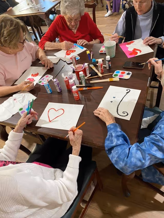 A group of elderly individuals seated around a wooden table engaged in a painting activity. They are using paintbrushes and various colors of paint to create artwork on white paper. The table is scattered with paint bottles, brushes, and paint palettes. The setting appears to be a communal indoor space with wooden flooring.