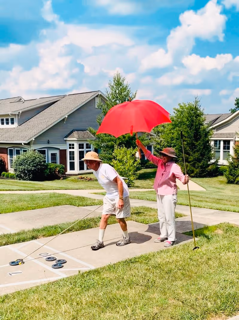 Two elderly people playing shuffleboard outdoors on a sunny day. One person is bending to push a shuffleboard puck while the other holds a bright red umbrella and a shuffleboard cue stick. They are on a concrete shuffleboard court with grass and residential buildings in the background under a partly cloudy blue sky.