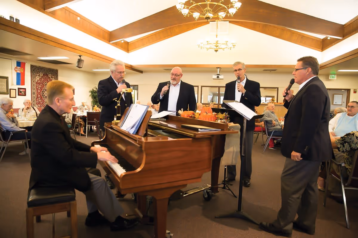 A pianist and four male singers perform by a grand piano as senior residents sit at tables in a community dining/activity room.