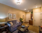 Cozy living room with a gray sofa adorned with patterned pillows, a wooden coffee table with decorative items, a side table with a lamp, two framed botanical prints on the wall, and a small entryway area with a door and a cabinet.