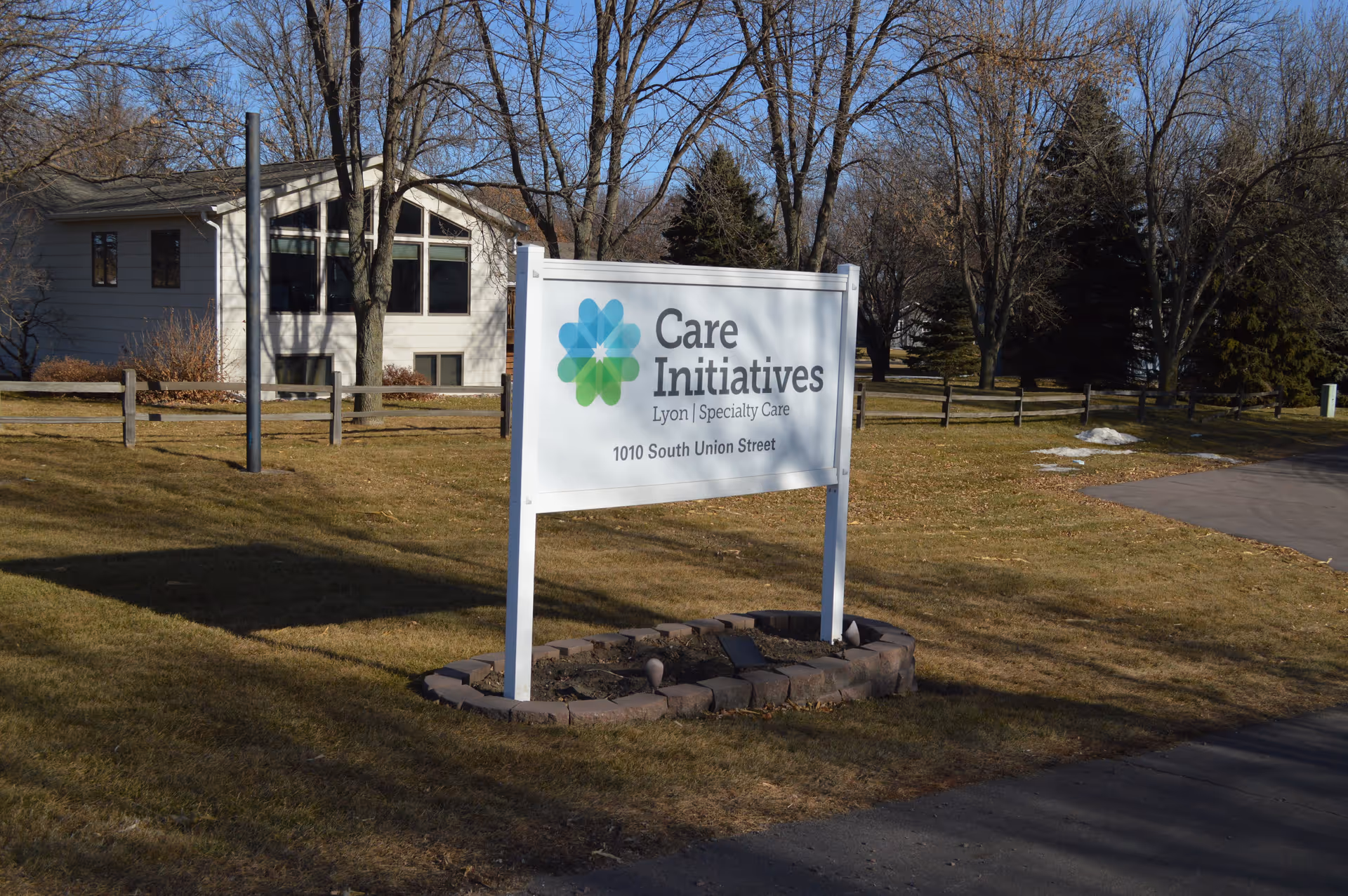 Outdoor view of a Care Initiatives Lyon Specialty Care facility sign on a grassy area with a house and trees in the background under a clear blue sky.