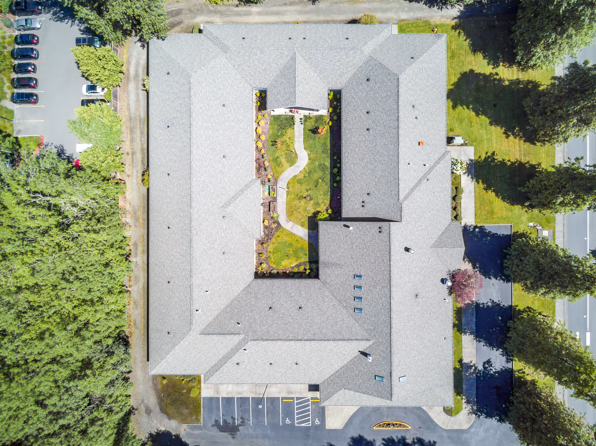 Aerial view of Creekside Place Memory Care building showing a U-shaped roof with a landscaped courtyard in the center featuring a winding pathway and greenery. Surrounding the building are parking areas, trees, and grassy spaces.