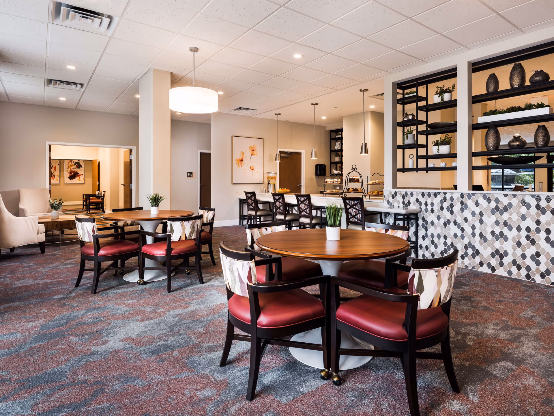 Bright communal dining and seating area with round tables, red-upholstered chairs, and a serving counter in a senior living facility.