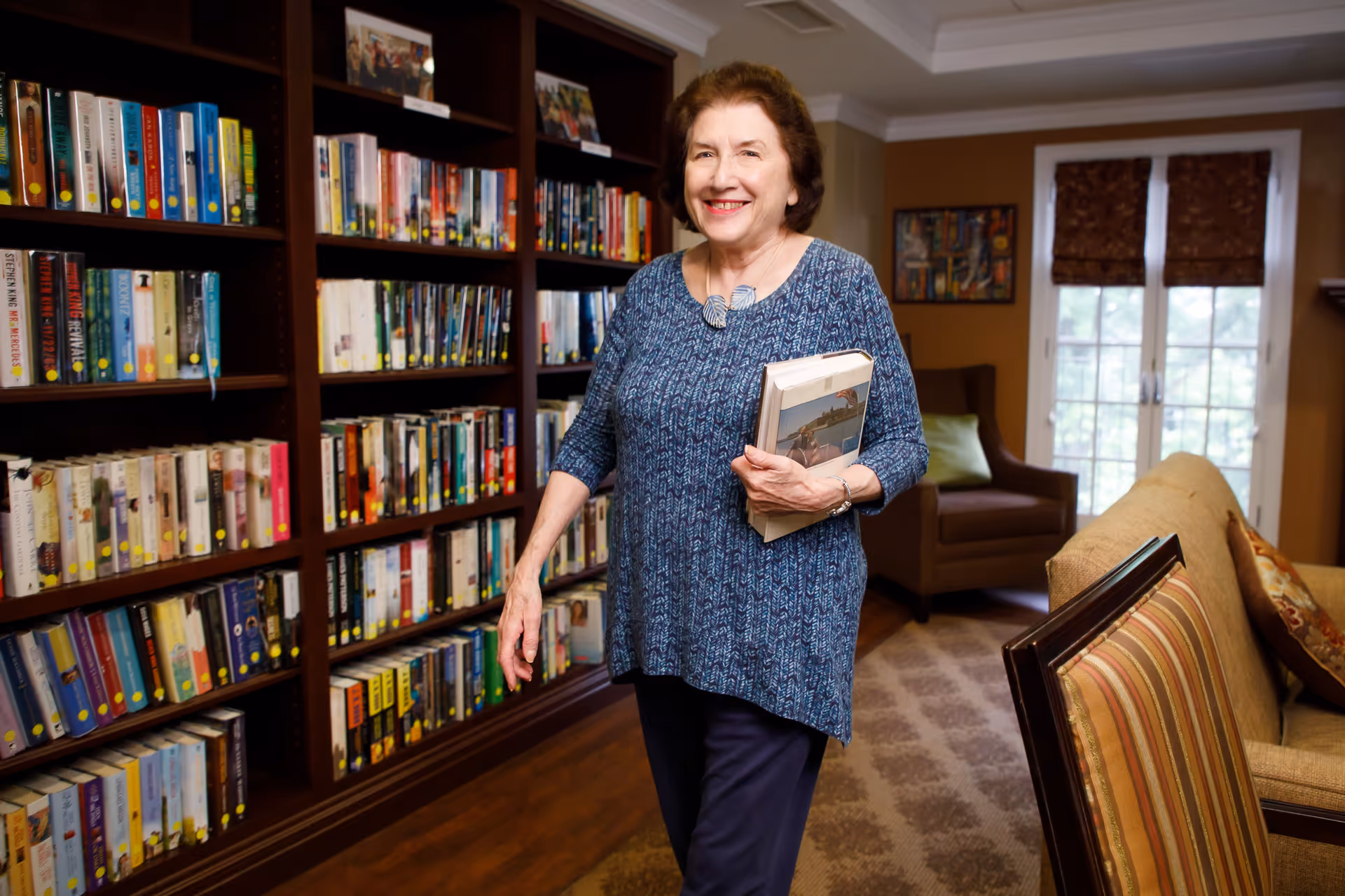 An elderly woman smiling and holding books while standing in a cozy room with a large bookshelf filled with books, a comfortable armchair, a sofa, and a window with curtains in the background.
