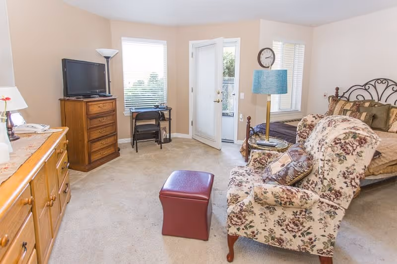 Furnished bedroom with a bed, floral armchair and ottoman, wooden dressers, a TV, and a door leading outside.
