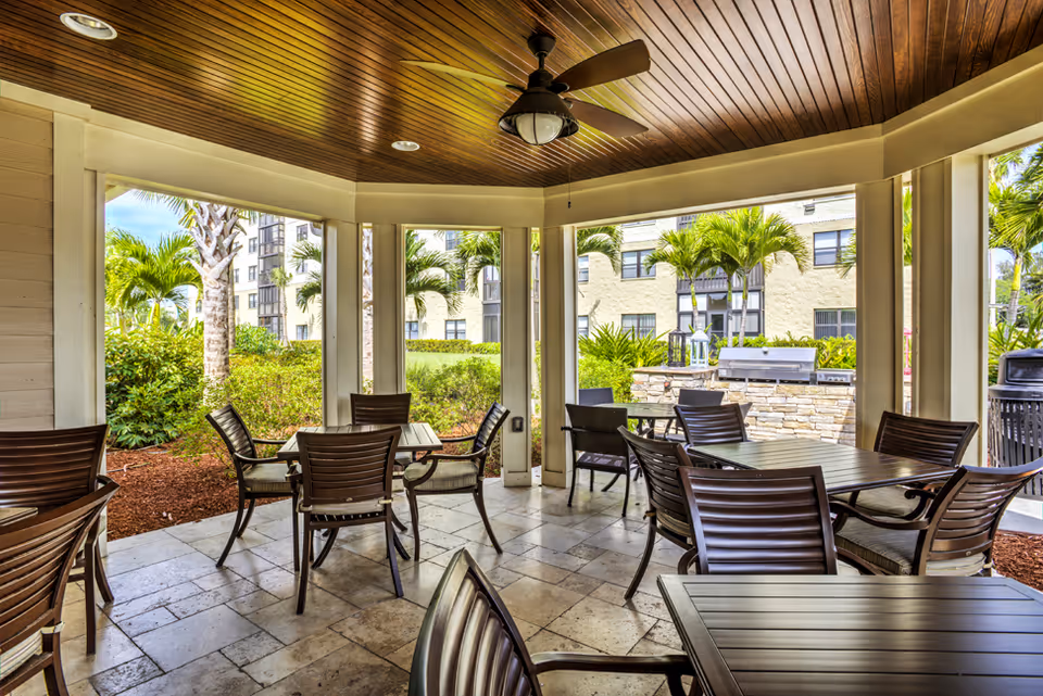 Covered outdoor seating area with multiple tables and chairs under a wooden ceiling with a ceiling fan. The area is surrounded by open windows showing palm trees, bushes, and a building in the background. There is a stone grill or barbecue station visible outside.