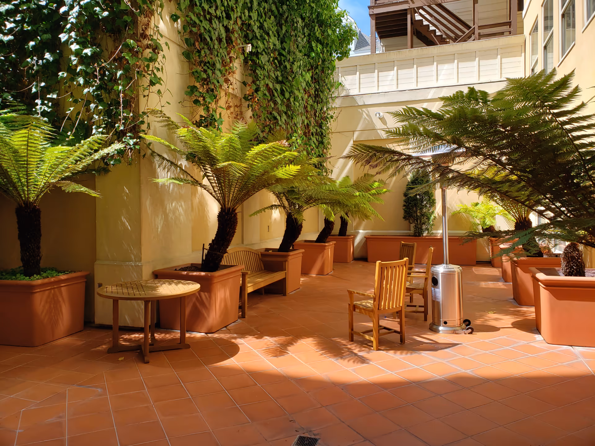 Outdoor patio area with terracotta tiled floor, large potted ferns, wooden chairs and a bench, and a tall outdoor heater. The space is enclosed by light-colored walls with some climbing plants and windows on one side.