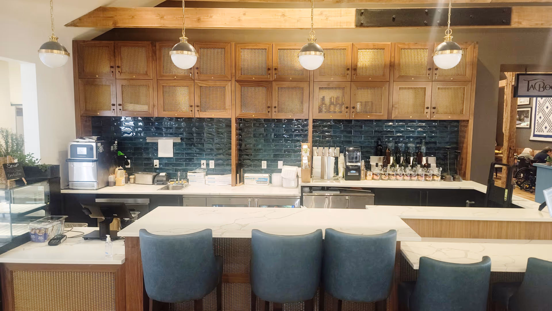Interior view of a modern kitchen area with a white marble countertop island and four blue upholstered chairs. The back wall features wooden cabinets with a dark green tiled backsplash. Various kitchen appliances and coffee dispensers are visible on the counter. Three pendant lights hang from the ceiling above the island.