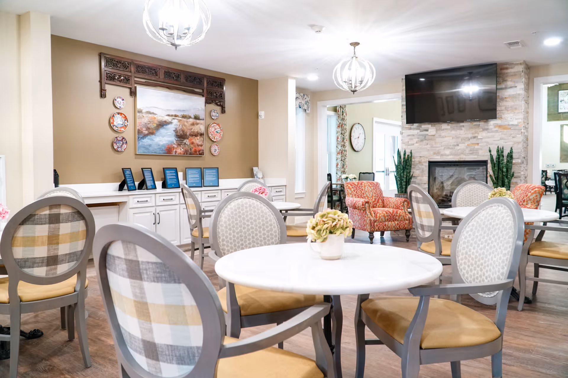 A bright and inviting common area with round tables and cushioned chairs arranged for seating. The room features a stone fireplace with a mounted flat-screen TV above it, two patterned armchairs, and decorative plants on either side of the fireplace. The walls are adorned with a large painting and decorative plates, and there are modern light fixtures hanging from the ceiling.