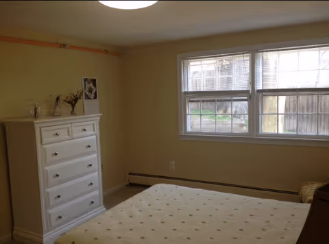 A bedroom with beige walls and carpet, featuring a white chest of drawers with decorative items on top, a bed with a light-colored polka dot bedspread, and a window with white blinds letting in natural light.