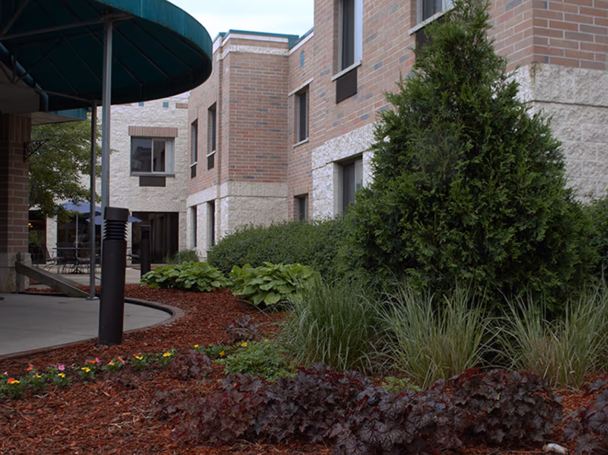 Outdoor garden area with various plants and shrubs in front of a brick and stone building. There is a covered walkway with a green canopy and outdoor seating visible in the background.