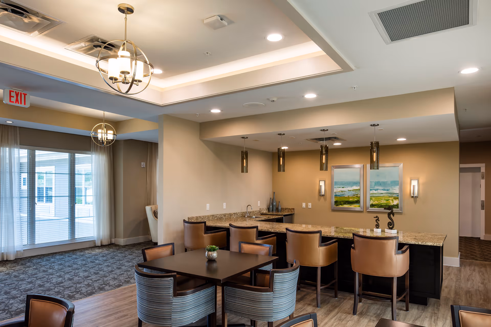 A modern common area with a granite countertop bar featuring five tan leather chairs. In front of the bar is a dark wood table with four striped upholstered chairs. The room has beige walls, two framed landscape paintings, pendant lights above the bar, and a ceiling light fixture. Large windows with sheer curtains allow natural light into the space.