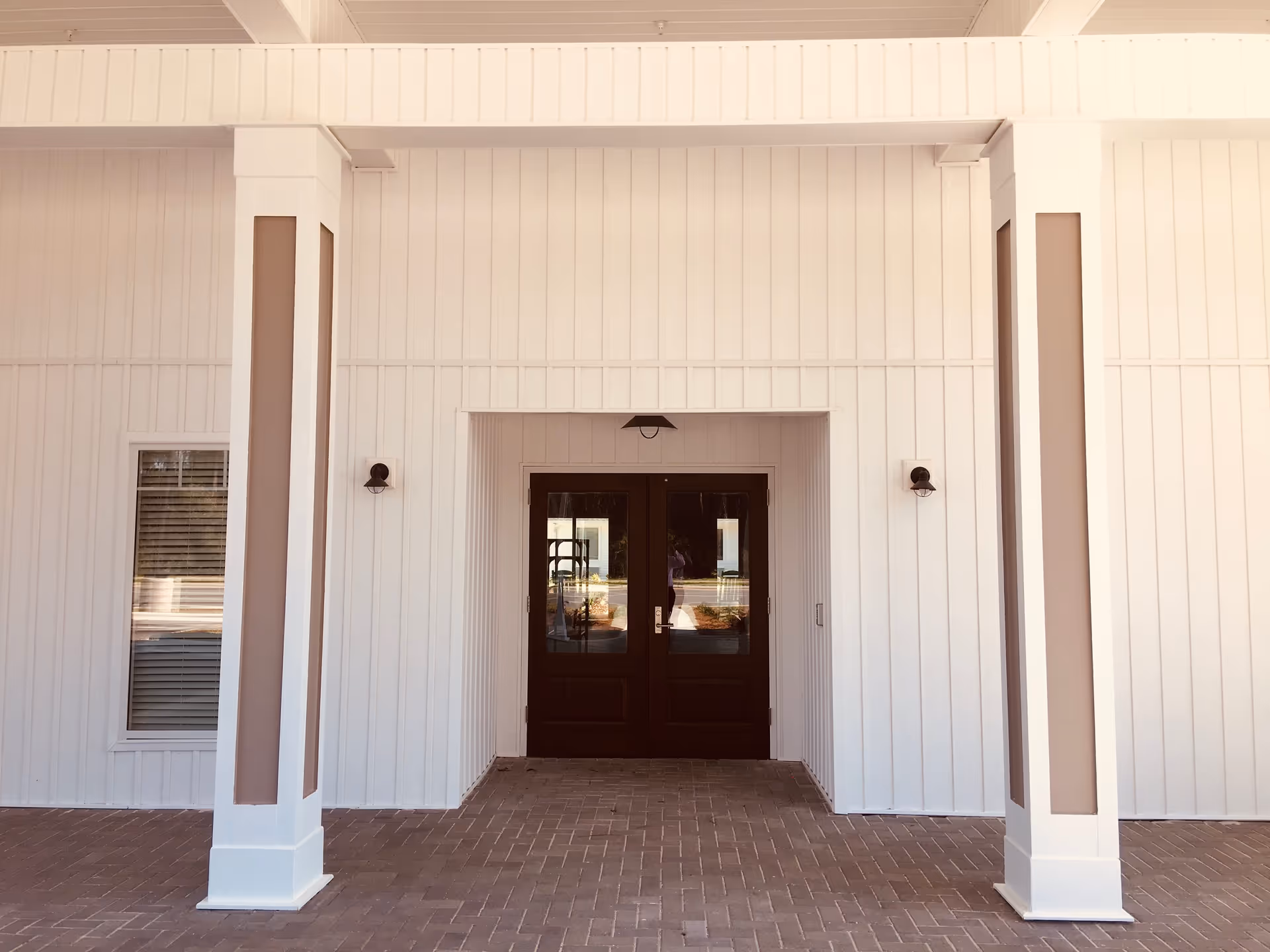 Entrance to a building with white paneled walls, two white columns with beige accents, a brick floor, and double dark wooden doors with glass panels. There are two wall-mounted light fixtures on either side of the doors and a ceiling light above the entrance.