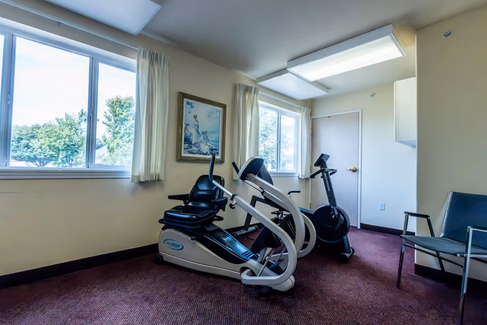 A small exercise room with two stationary exercise bikes positioned near two large windows with cream-colored curtains. There is a framed painting on the wall between the windows and a simple chair with metal arms to the right. The room has beige walls and a maroon carpet.