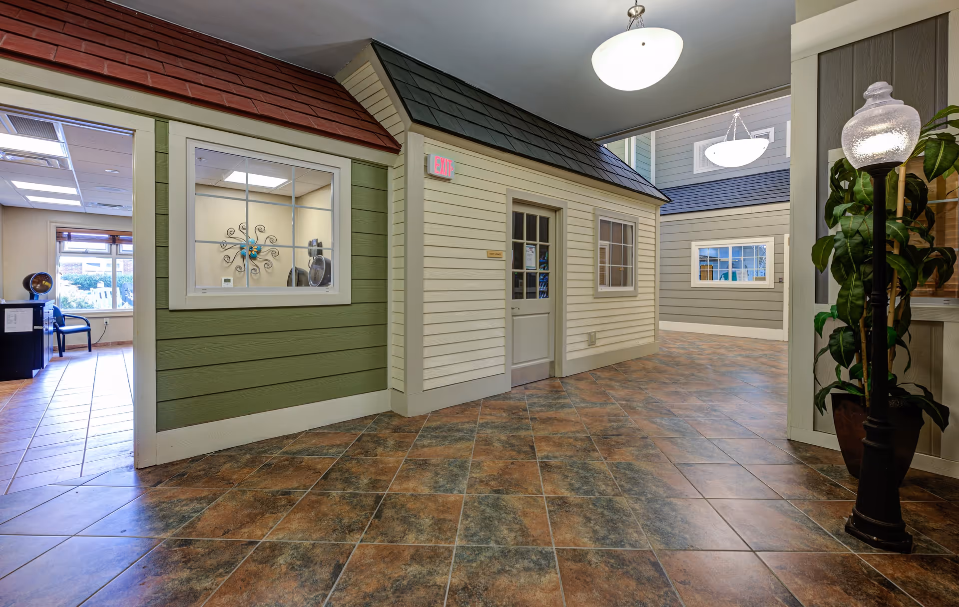 Interior hallway of a senior living facility designed to look like a small village street with house-like structures featuring windows and doors. The floor is tiled with a brown and gray pattern, and there are ceiling lights and a decorative street lamp with a plant beside it.