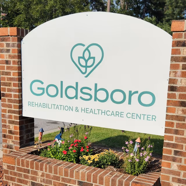 White entrance sign reading "Goldsboro Rehabilitation & Healthcare Center" mounted between brick pillars with flowers planted below.