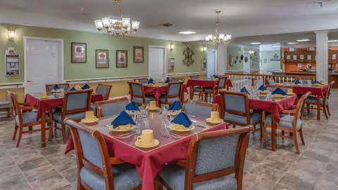 A dining room in a senior living facility with multiple tables covered in red tablecloths, each set with yellow cups, plates, glasses, and blue folded napkins. The room has green walls adorned with framed pictures and decorative wall art. There are chandeliers hanging from the ceiling providing lighting, and a counter area is visible in the background.
