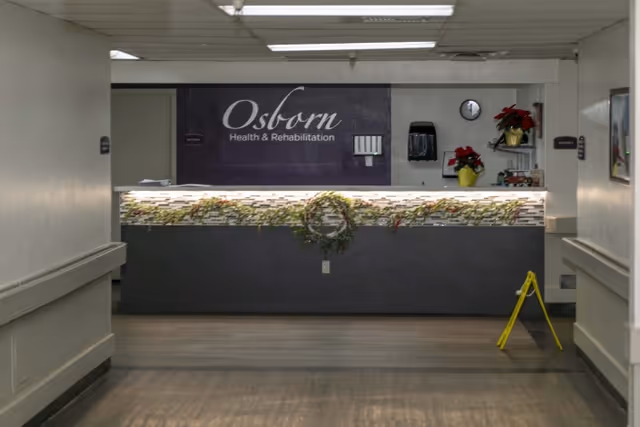 Reception desk area of Osborn Health & Rehabilitation with sign behind the counter, holiday plants, and a wet-floor sign in the hallway.