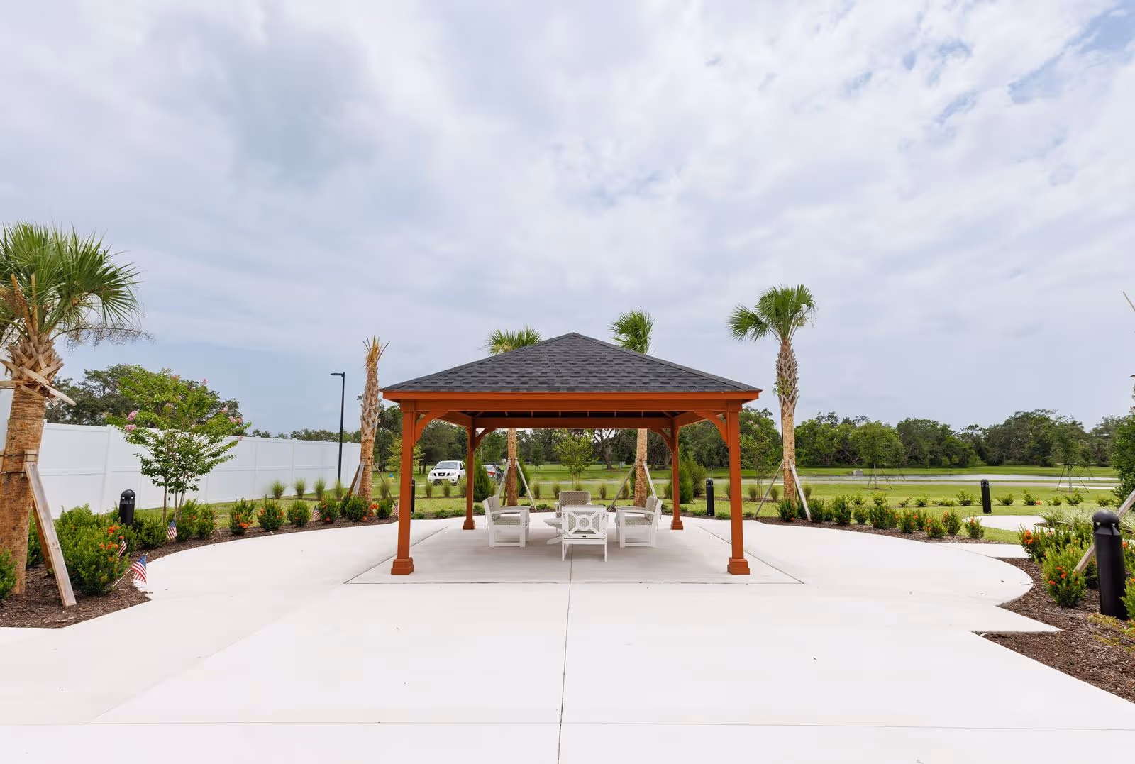 Outdoor seating area with a wooden gazebo structure and white chairs on a concrete patio surrounded by palm trees and greenery under a cloudy sky.
