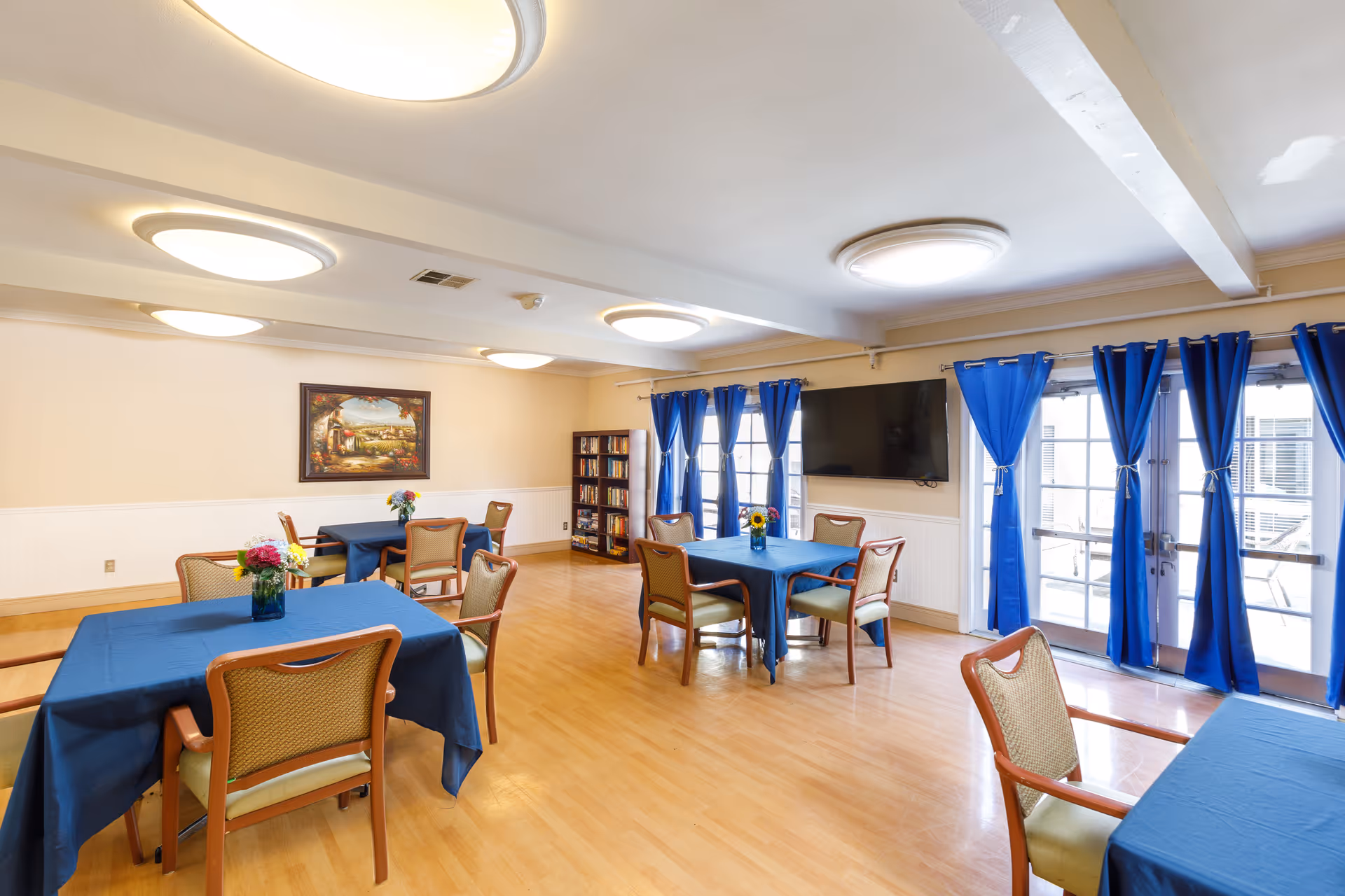 A bright, spacious common area with wooden floors and beige walls. The room has several tables covered with blue tablecloths, each surrounded by chairs with wooden frames and green cushions. Small flower arrangements are placed on the tables. Blue curtains frame the glass doors and windows on the right side, and a large flat-screen TV is mounted on the wall. A bookshelf filled with books is visible in the back corner, and a framed painting hangs on the far wall.