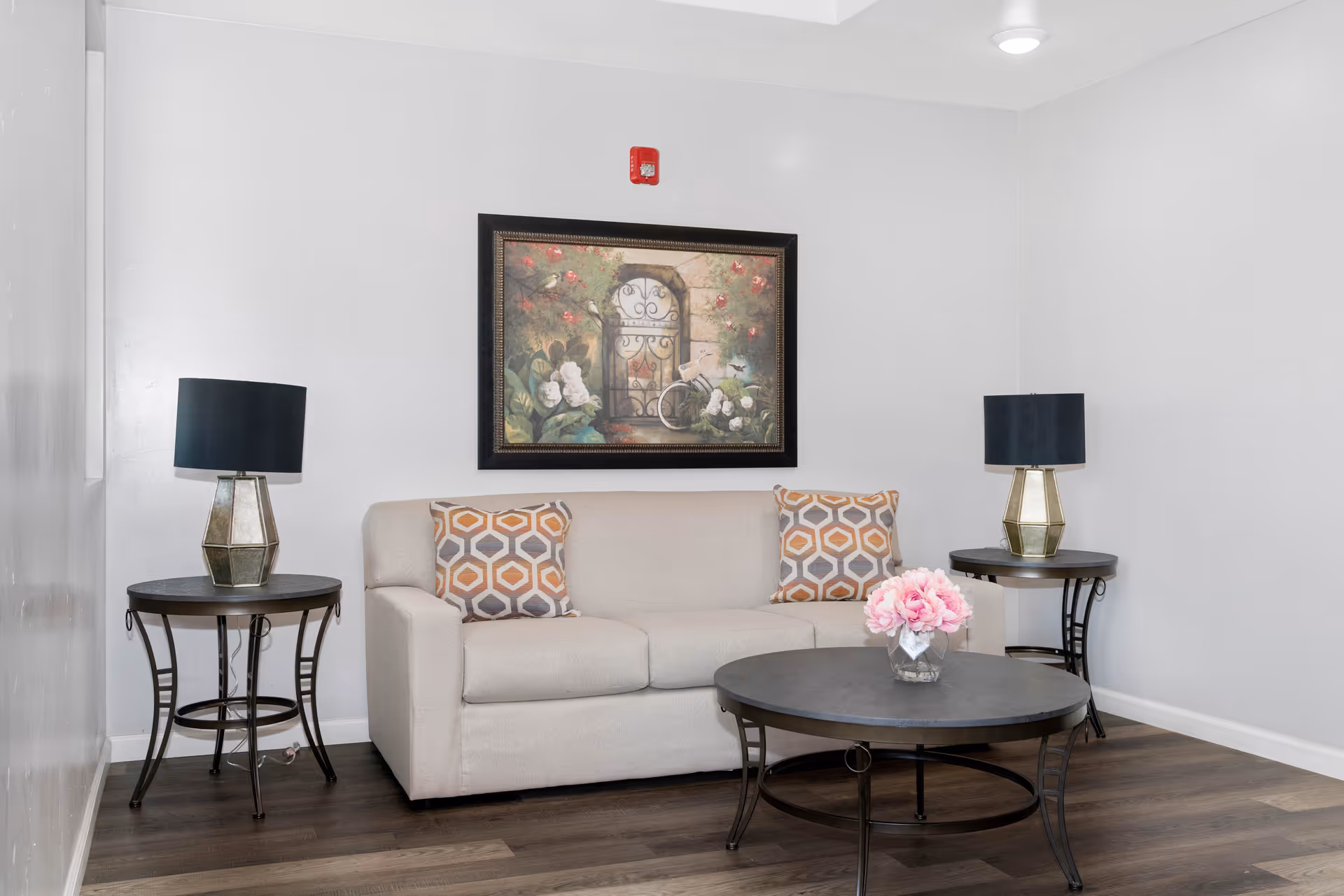 A small sitting area with a beige loveseat, round coffee table with pink flowers, two side tables with lamps, and framed artwork on a white wall.