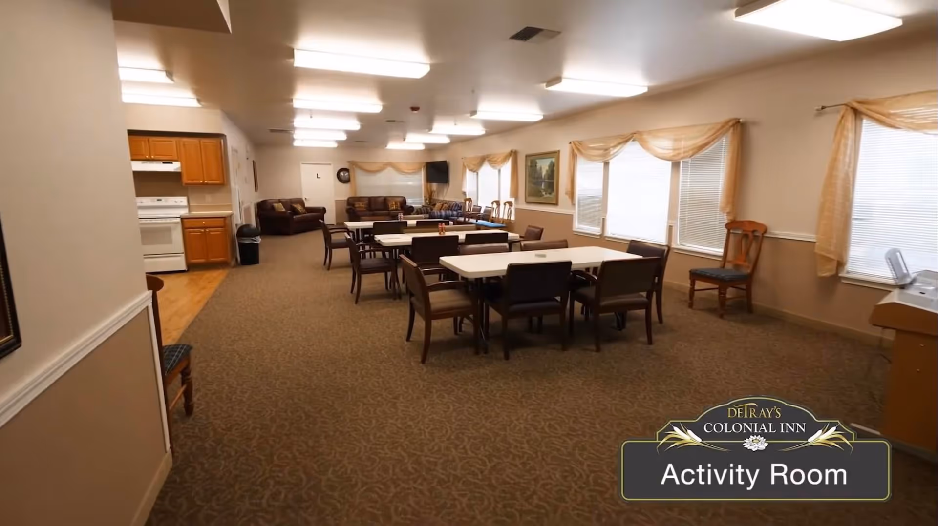 A spacious activity room with several tables and chairs arranged for group activities. The room has carpeted flooring, beige walls with white trim, and multiple windows with sheer curtains allowing natural light. There is a small kitchen area with wooden cabinets and a white stove on the left side. At the far end, there are sofas and a wall-mounted television. A sign in the bottom right corner reads 'DeTray's Colonial Inn Activity Room'.