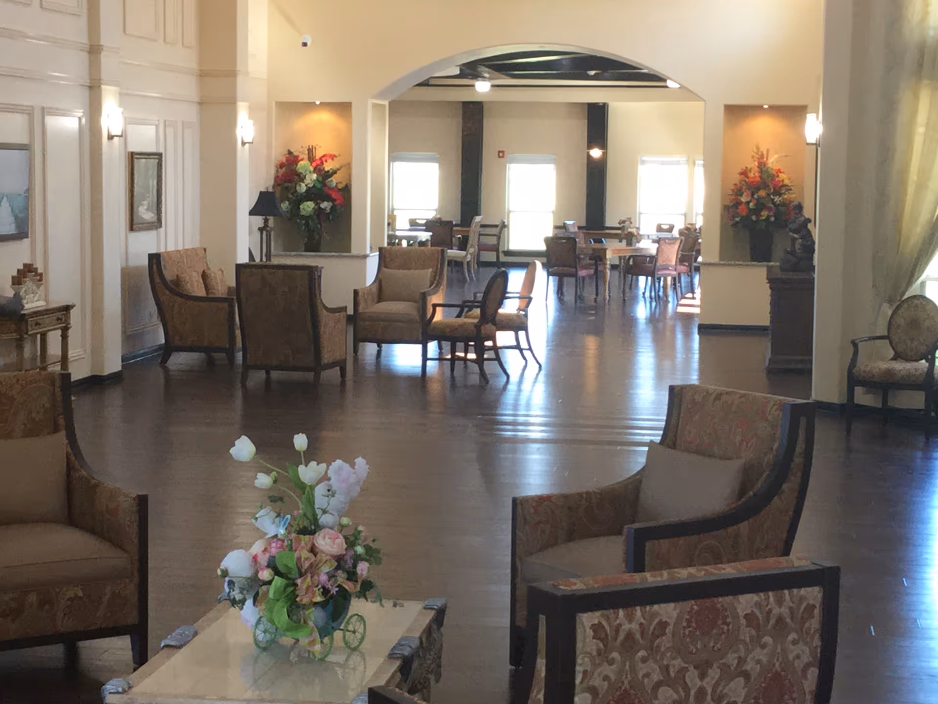 Spacious assisted living common area with armchairs, floral centerpieces, and a dining area visible in the background.