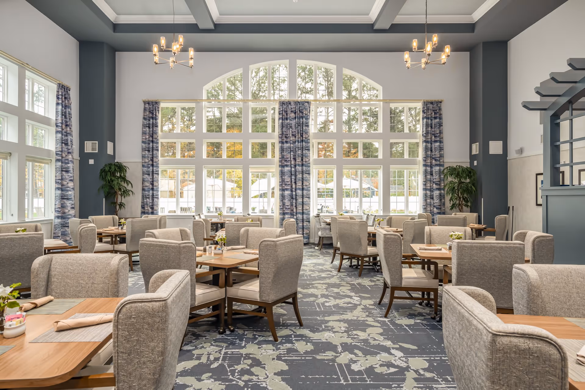 A spacious dining room with multiple wooden tables and cushioned chairs arranged neatly. Large windows with blue patterned curtains allow natural light to fill the room. The ceiling has decorative beams and two modern chandeliers. There are plants placed near the walls, and the carpet has a blue and gray abstract pattern.