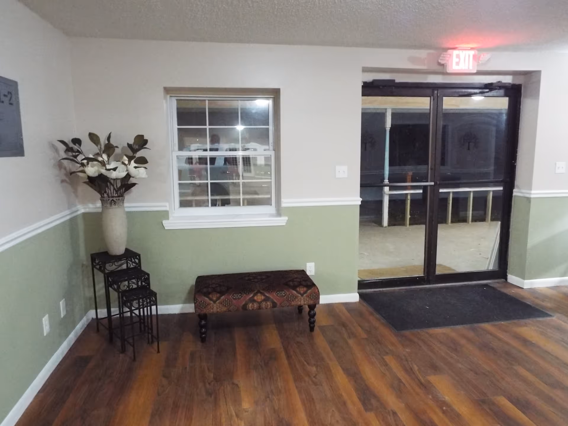 Small entry lobby with wood-look floor, a patterned bench, a tall vase on nested stands, a window and glass double doors with an illuminated EXIT sign.