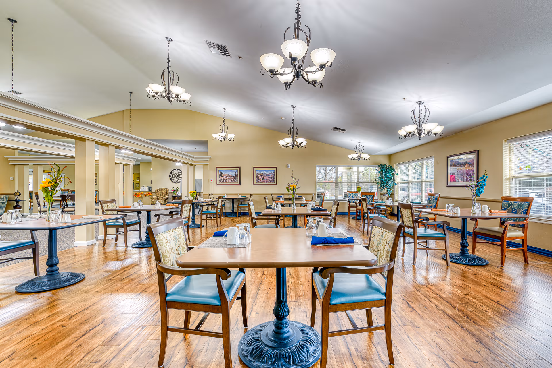 Spacious assisted-living dining room with neatly arranged tables and chairs, chandeliers, and large windows.