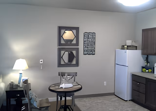 A small dining area and kitchen corner in a senior living facility. The dining area has a round glass-top table with two chairs. On the wall above the table are two decorative mirrors and a piece of wall art. To the right is a white refrigerator next to wooden kitchen cabinets and a countertop. A lit table lamp is on a small dark side table to the left.