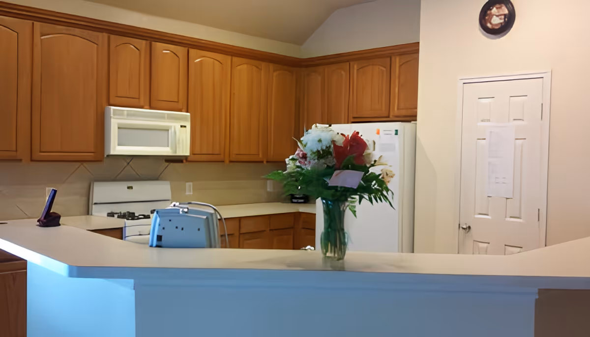 Interior view of a kitchen with wooden cabinets, a white stove with a microwave above it, a white refrigerator, and a countertop with a vase of flowers and a toaster on it. There is a white door with papers attached to it and a decorative plate hanging on the wall above the door.