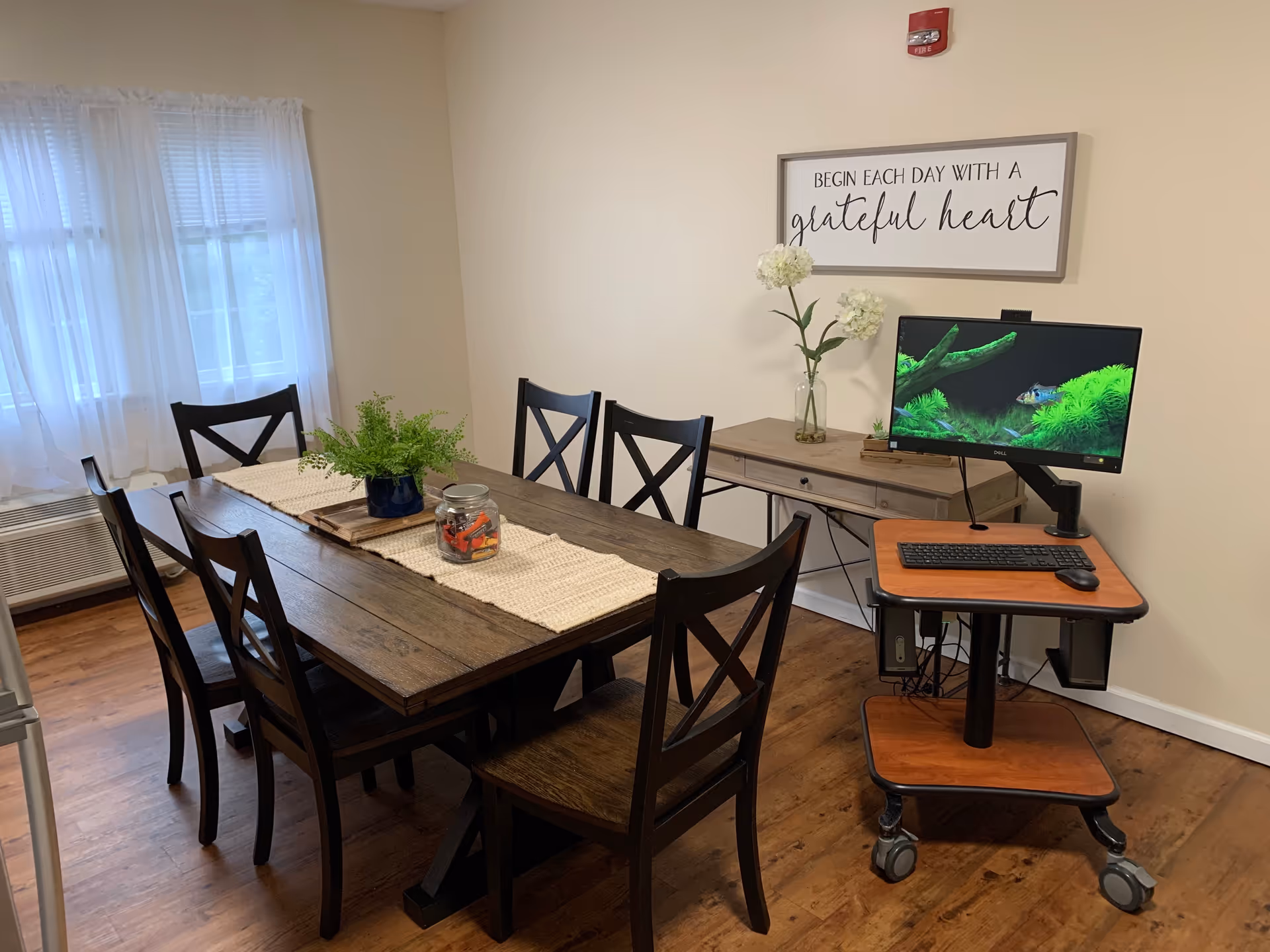 A dining room with a wooden table and six matching chairs. The table has a beige runner, a small potted fern, and a jar with candy. There is a window with sheer white curtains on the left wall. Against the right wall is a wooden console table with a vase holding white flowers and a framed sign above it that reads 'BEGIN EACH DAY WITH A grateful heart'. In front of the console table is a mobile computer workstation with a monitor displaying an aquarium screensaver, a keyboard, and a mouse. The floor is wood, and the walls are painted light beige.