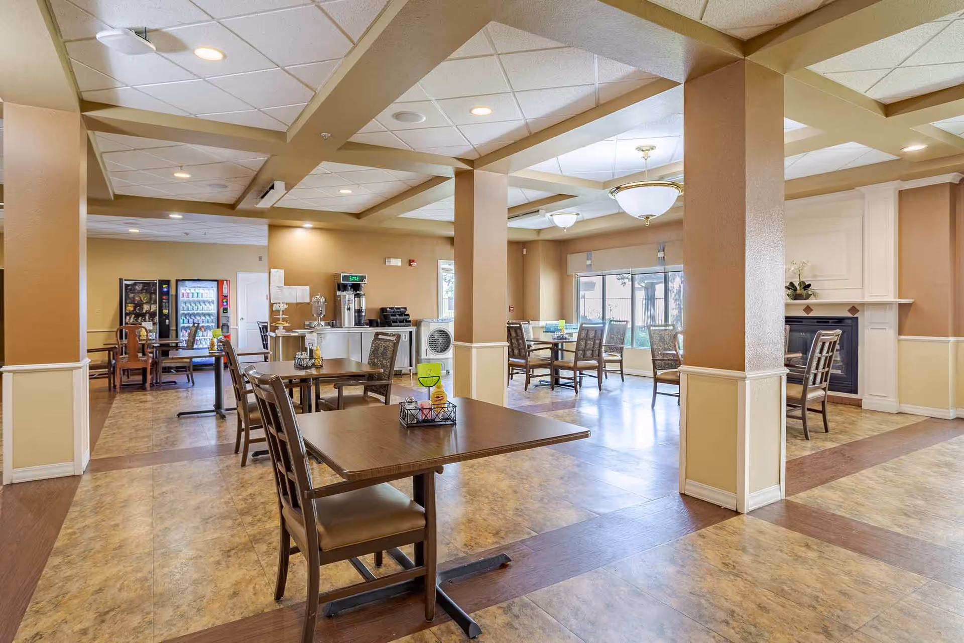 A spacious dining area in an assisted living facility with several wooden tables and chairs arranged neatly. The room features beige walls with white trim, a tiled floor with wood accents, large windows letting in natural light, and ceiling lights. In the background, there is a beverage vending machine and a coffee station. A fireplace with a white mantle is visible on the right side.