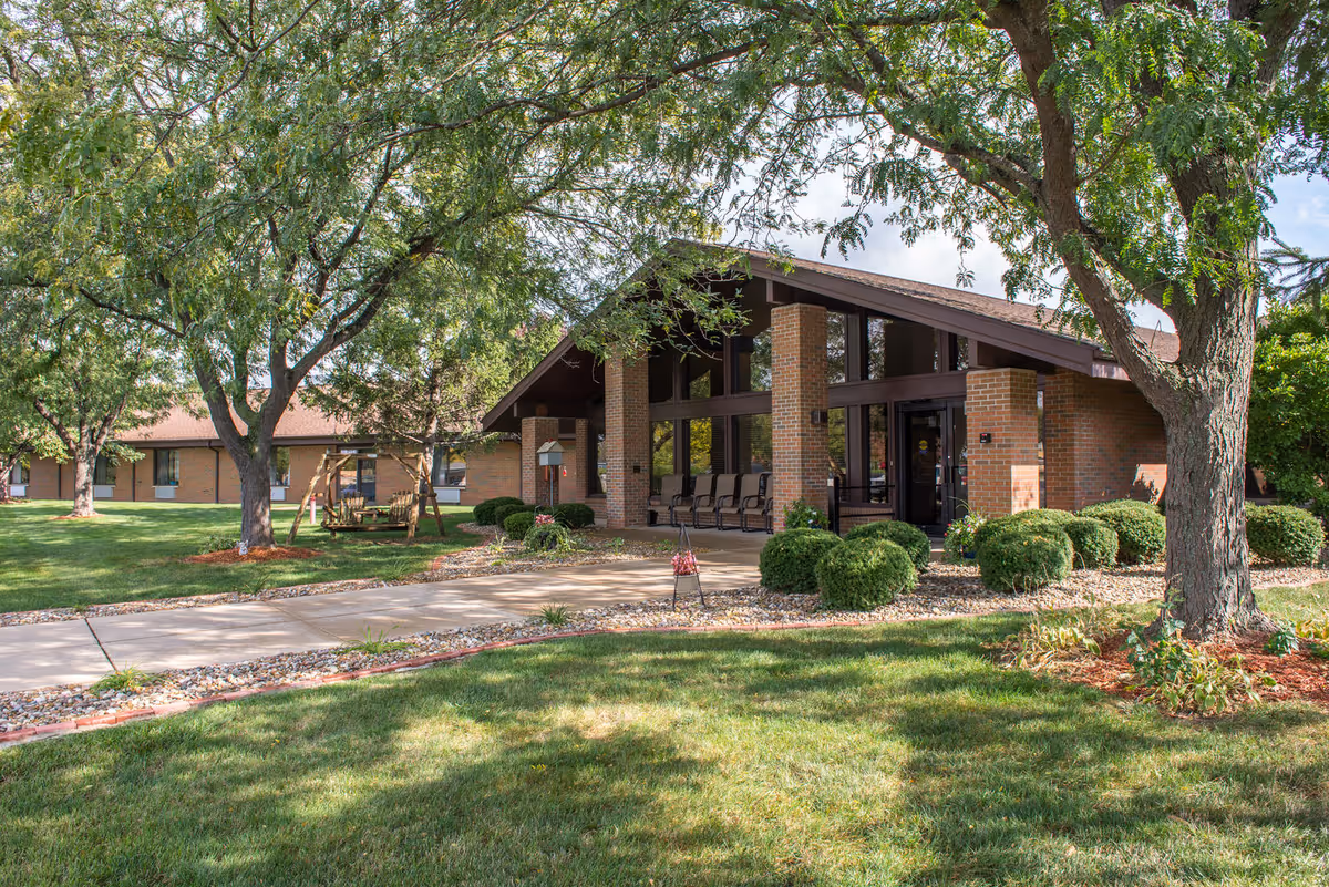 Exterior view of The Loft Rehabilitation and Nursing of Canton building with a brick facade, large windows, and a covered entrance. The surrounding area features green grass, trees, bushes, a paved walkway, and a wooden swing set.