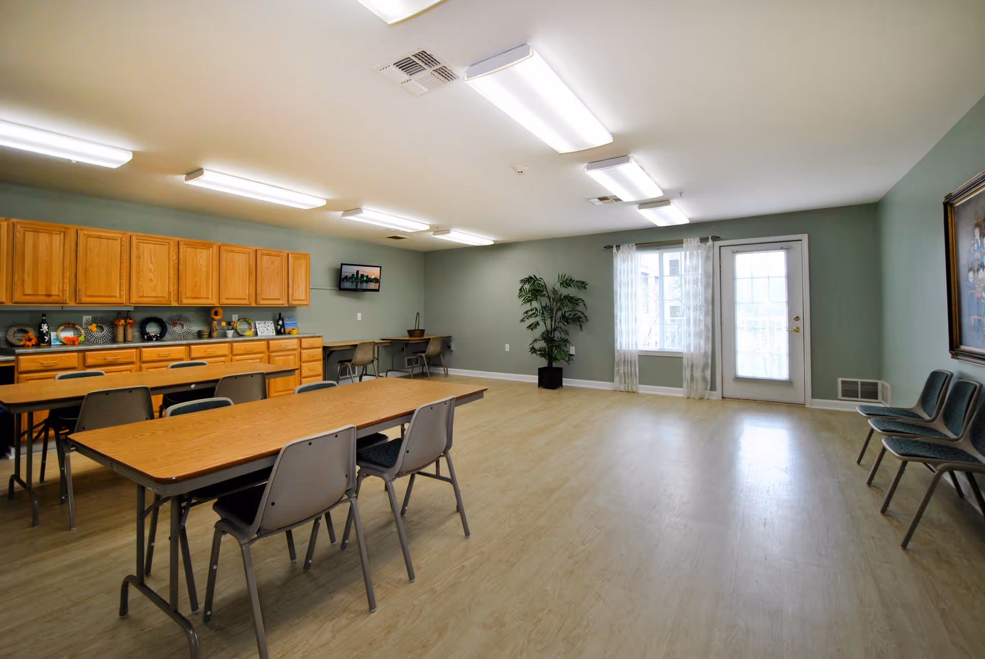 A spacious room with light green walls and wood flooring featuring two long tables with chairs, wooden cabinets along one wall, a small TV mounted on the wall, a potted plant near a window with sheer curtains, and a door with glass panels. Additional chairs are lined up against the opposite wall.