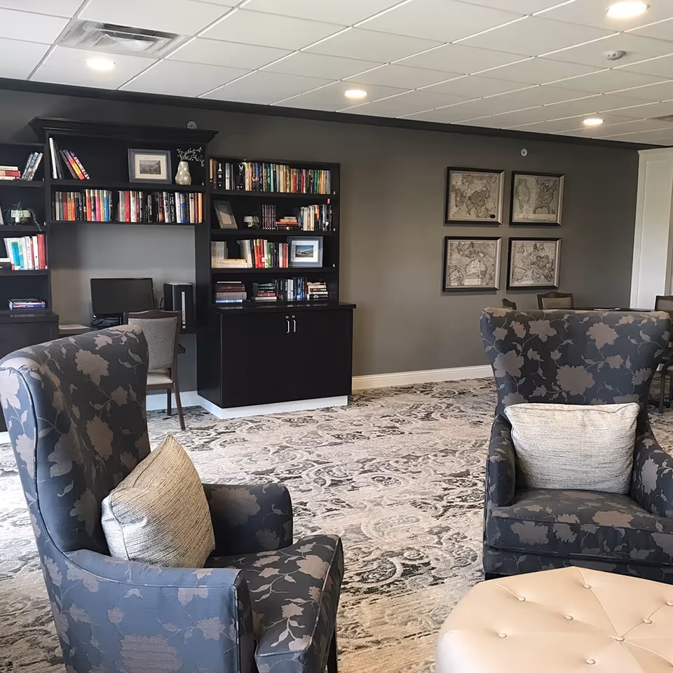 A cozy senior living common area with two patterned armchairs each with a light-colored pillow, a round tufted ottoman, a large bookshelf filled with books and decorative items, a computer workstation, and four framed vintage maps on the wall. The room has a patterned carpet and recessed ceiling lights.