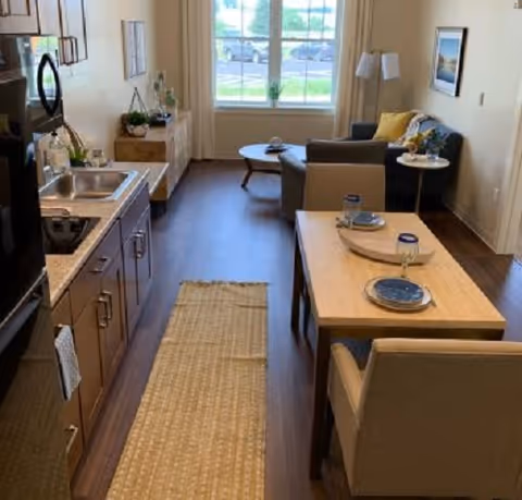 Open-plan assisted living unit with a kitchenette on the left, a dining table in the foreground, and a seating area by a large window.