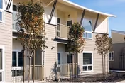 Exterior view of Casa Grande Senior Apartments showing a two-story building with beige siding, white trim, and a balcony on the upper floor. There are three young trees planted in front of the building and a clear sky above.