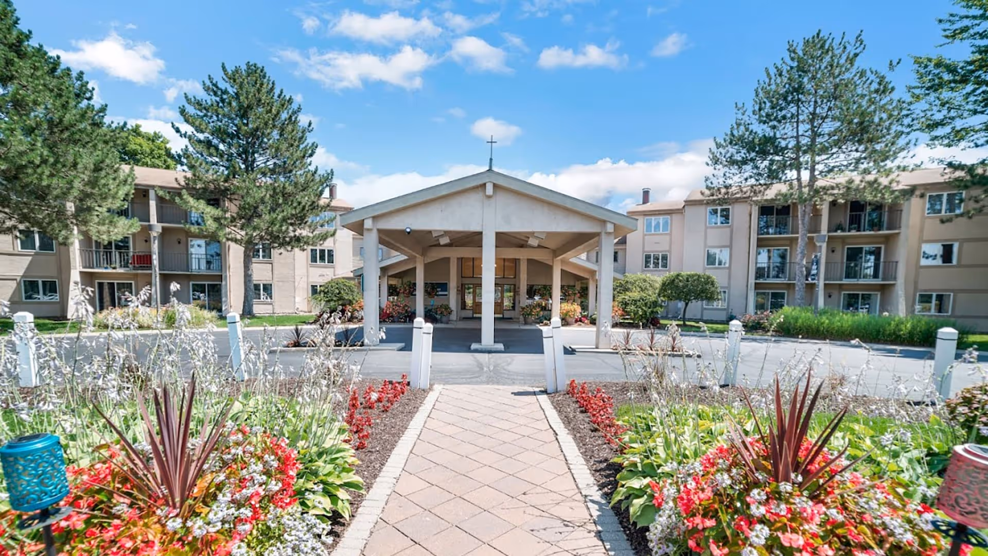 View of the entrance to a senior living facility with a covered drop-off area, surrounded by well-maintained flower beds and greenery, with multi-story residential buildings in the background under a blue sky with scattered clouds.