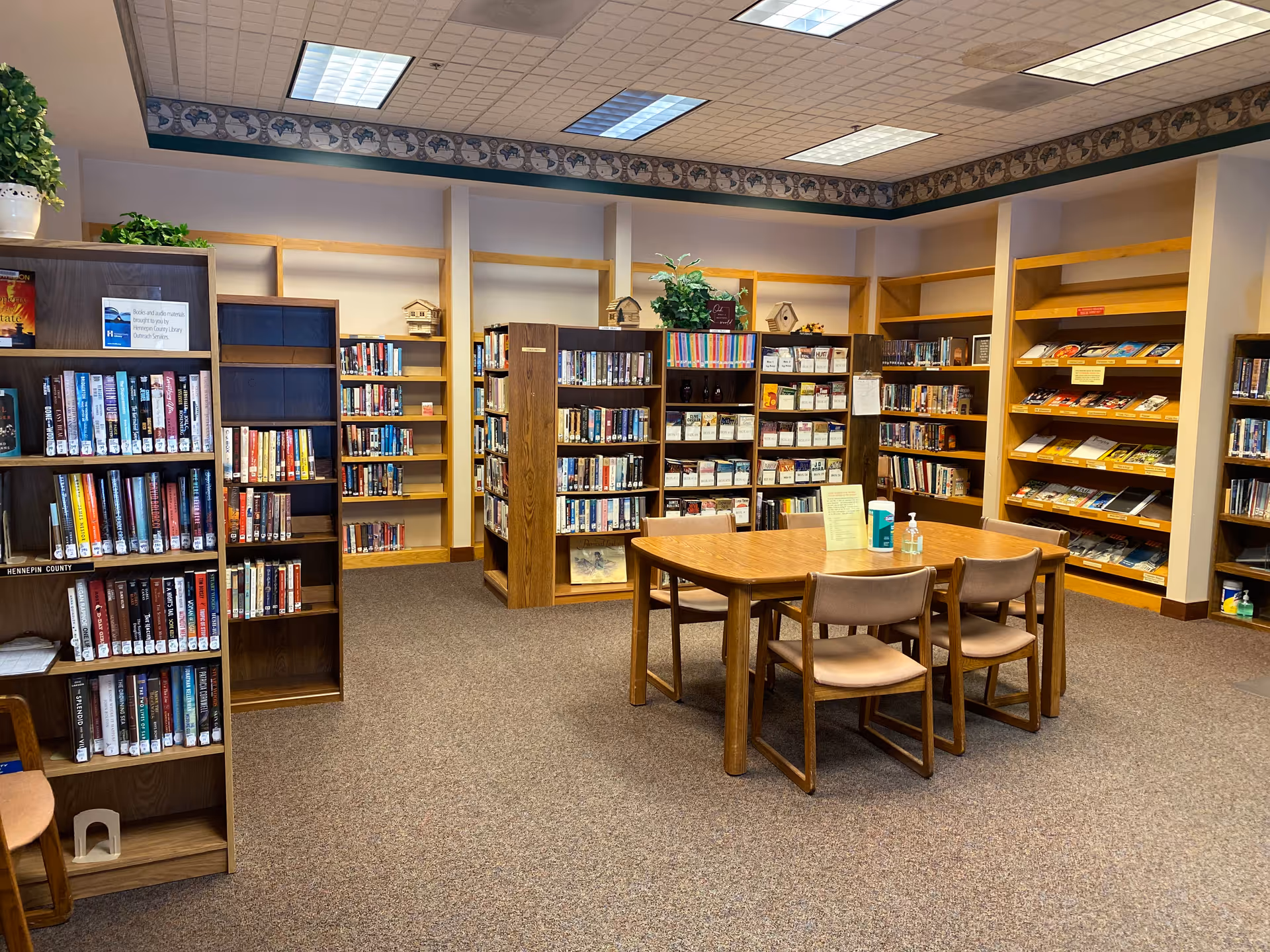 Interior view of a library room with multiple wooden bookshelves filled with books. A wooden table with four chairs is placed in the center, with hand sanitizer and a small sign on the table. The room has carpeted flooring and fluorescent ceiling lights.