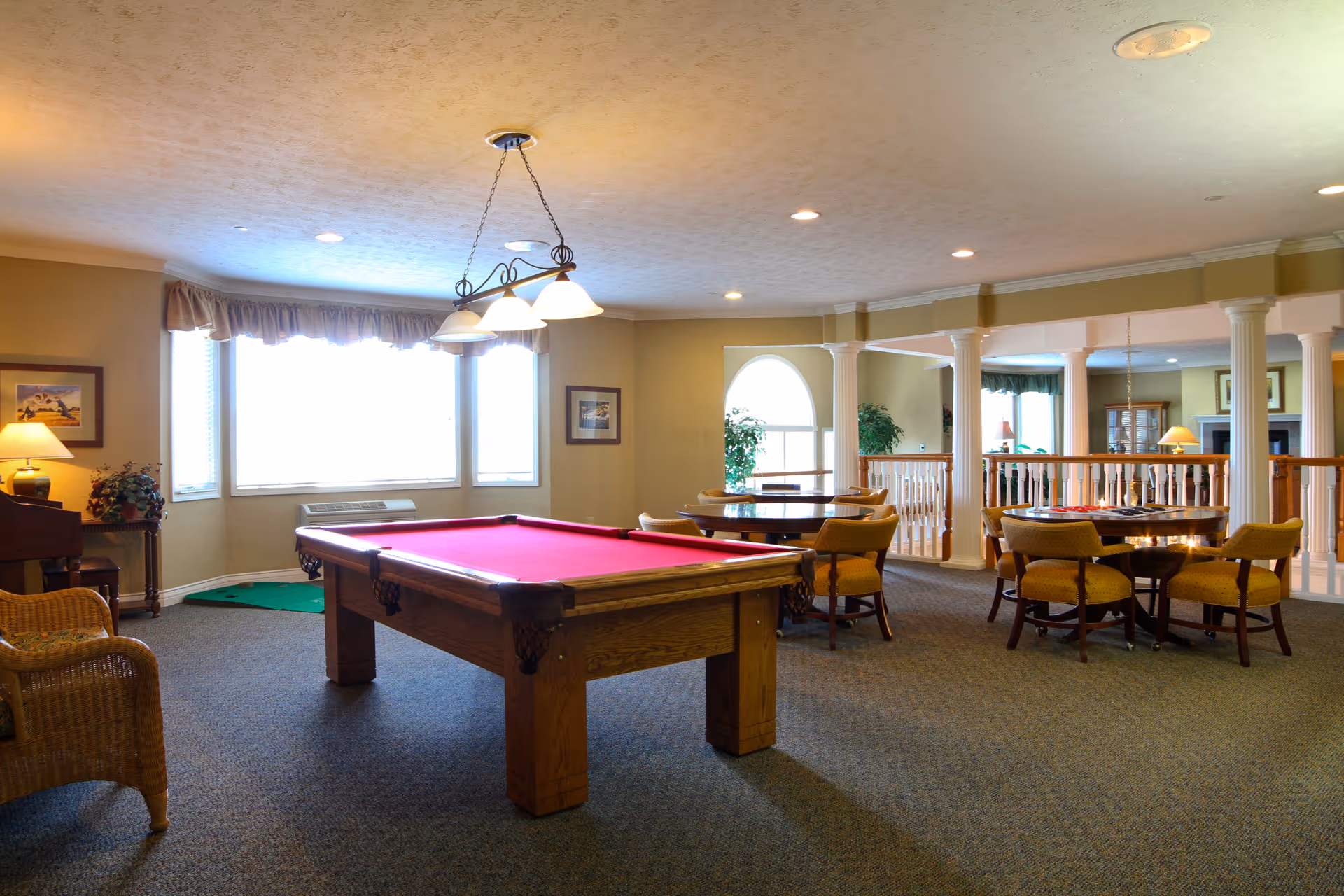 A spacious common area in a senior living facility featuring a red felt pool table in the center, surrounded by carpeted flooring. To the right, there are round tables with yellow cushioned chairs arranged for socializing or games. The room is well-lit with ceiling lights and a hanging light fixture above the pool table. Large windows with valances allow natural light to fill the space. Decorative columns and railings separate this area from an adjacent room with more seating and a fireplace.